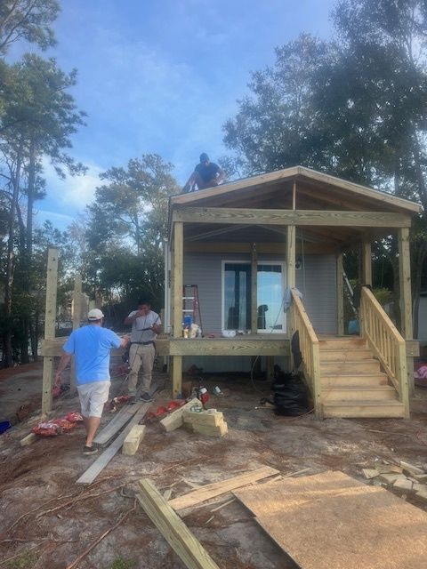 Construction workers building a small house with a porch and stairs. Blue sky, trees in the background.