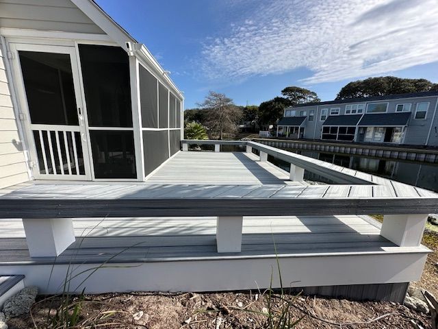 Deck with white railing, leading to a screened porch. Gray deck boards with water in the background.