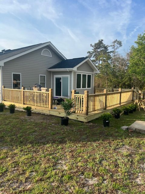 A newly constructed wooden deck extends from a gray house, surrounded by bushes in pots and a grassy yard.
