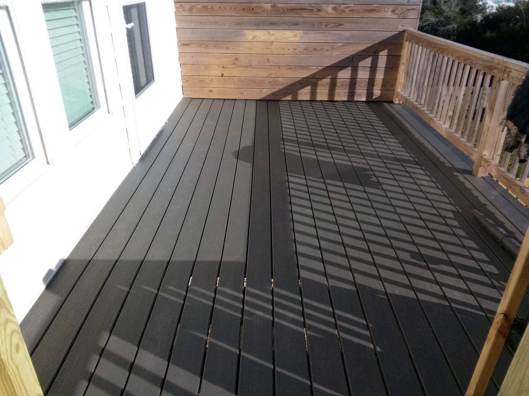 Dark gray deck with wooden railing and wall attached to a white house, in daylight.