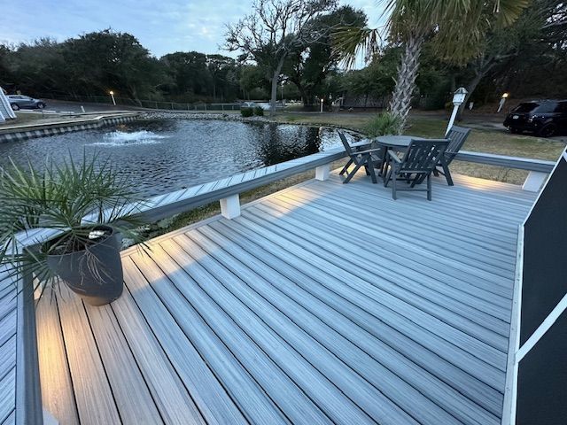 Deck with gray wooden planks overlooks a pond with a fountain. Furniture sits on the deck.