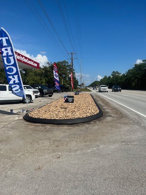 A car dealership with flags and cars, a median with rocks, and a road with cars on a sunny day.