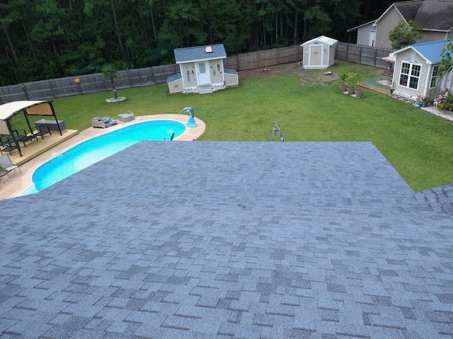 Overhead view of a backyard with a pool, sheds, and a house with a gray shingle roof.