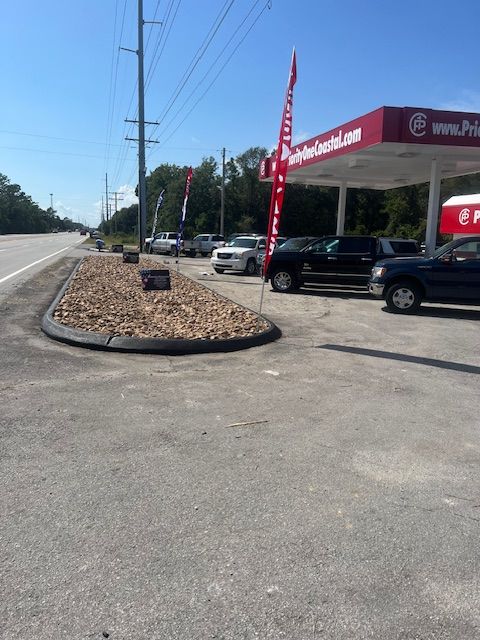Car dealership with gas station, cars parked on lot, flags waving, sunny day.