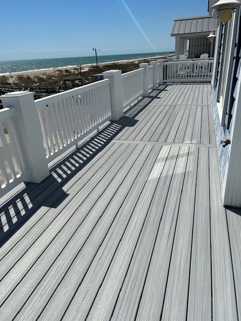 A wooden deck with white railing faces the beach. The deck boards are gray; the sky is blue.
