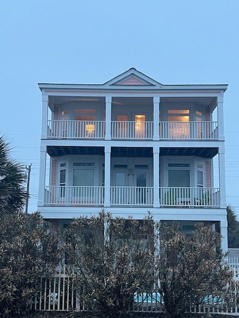 Three-story light pink house with balconies, white railing, and bay windows. The house sits behind a line of shrubs.