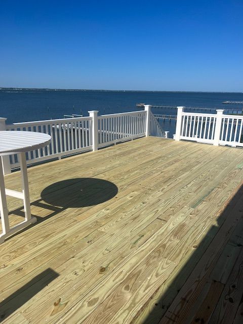 Wooden deck with white railing and table overlooking a body of water under a clear blue sky.