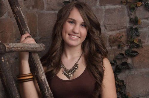 A young woman is holding a wooden ladder in front of a brick wall.