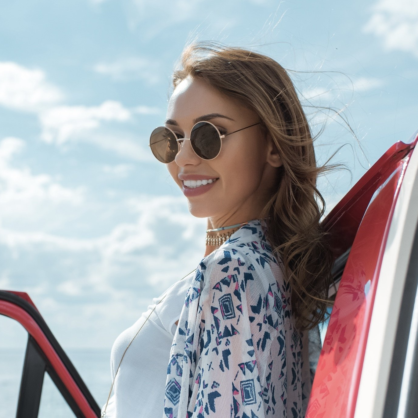 A woman wearing sunglasses is standing next to a red car.