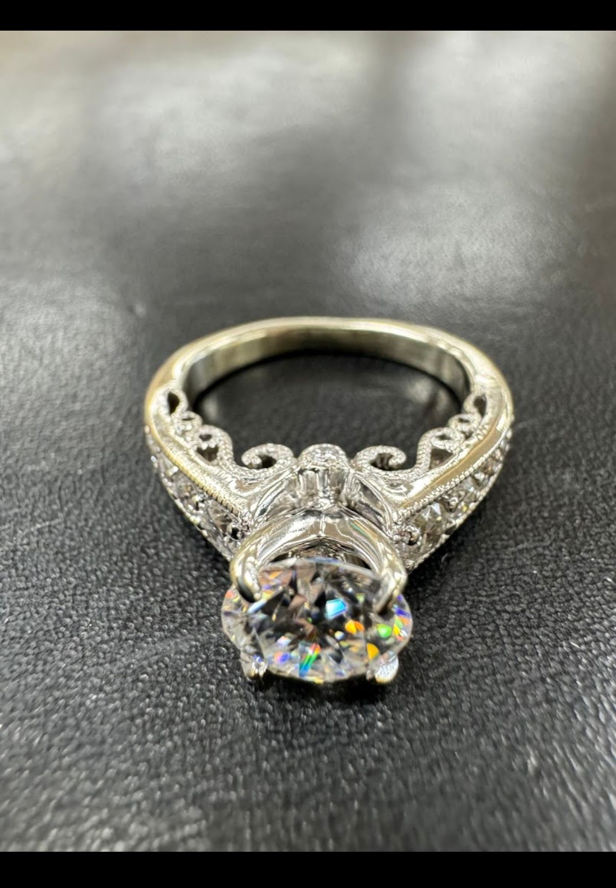 Ornate silver engagement ring with a large round diamond on a black surface