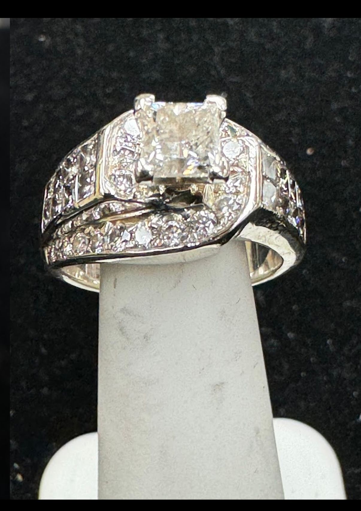 Diamond engagement ring with square center stone and pavé band on a white display stand