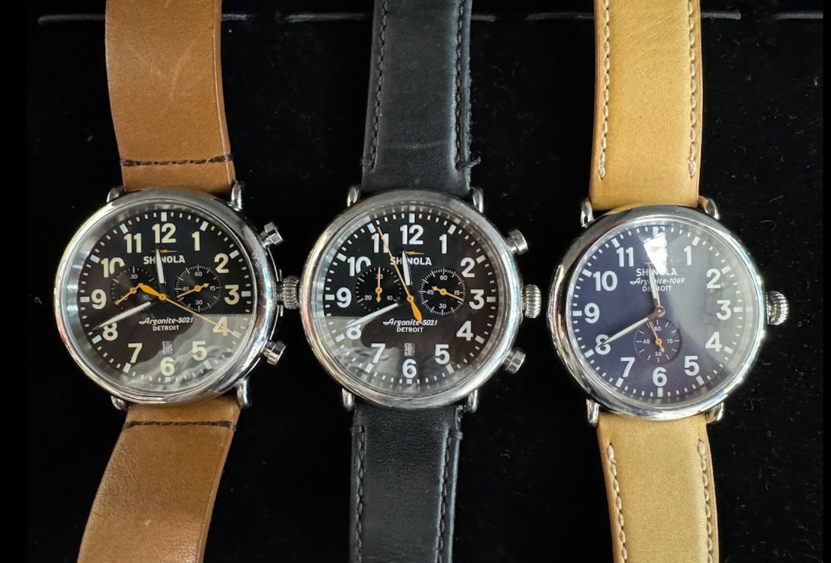 Three wristwatches with tan, black, and beige straps displayed side by side on a dark background