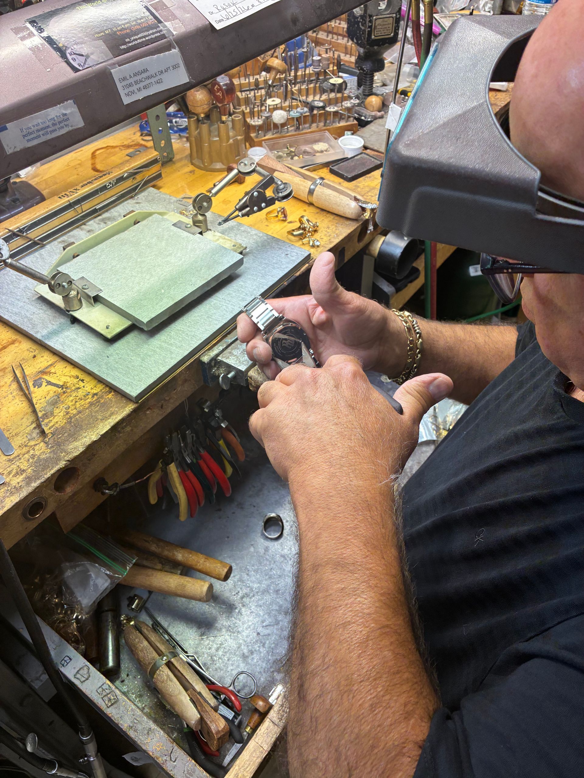 Person working at a cluttered jeweler’s bench, using tools on a small piece under a lamp.