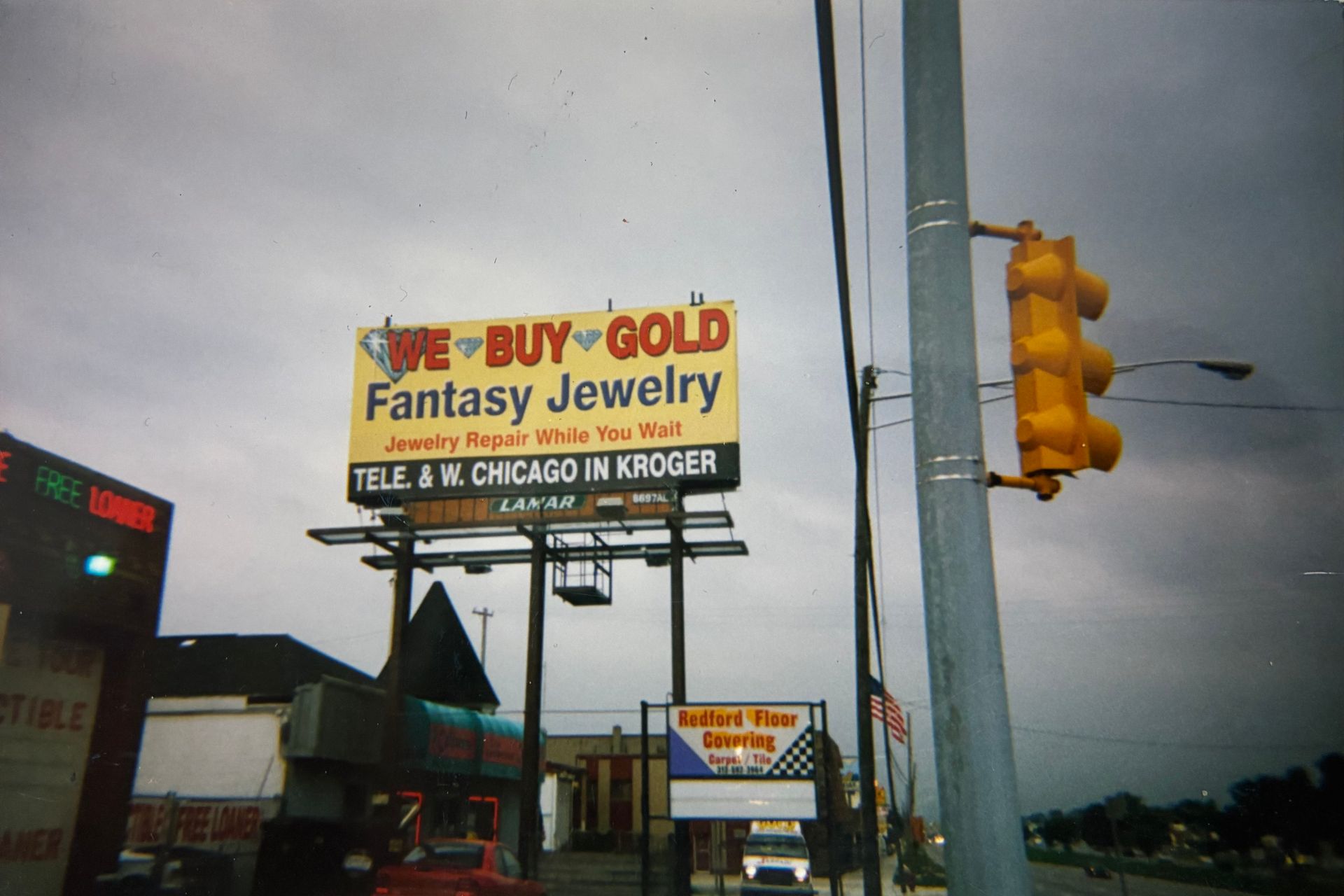 Street scene with a Fantasy Jewelry sign, roadside businesses, and a traffic light under a cloudy sky.