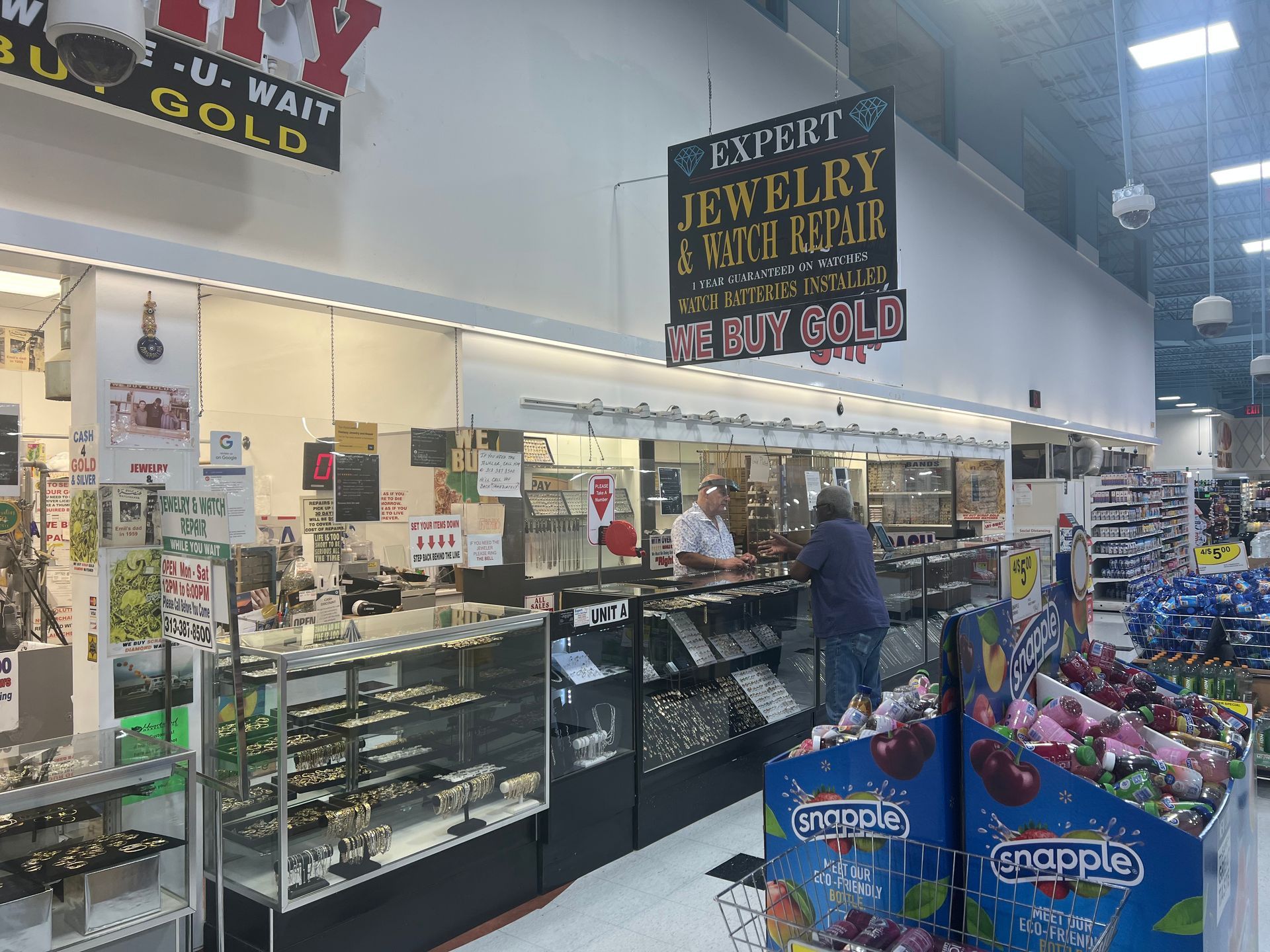 A jewelry repair kiosk inside a retail store with glass display cases, signs advertising gold buying, and service staff.