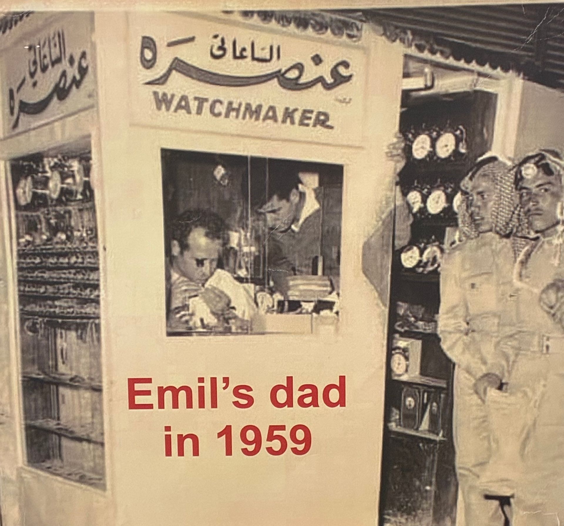 Vintage black-and-white shop scene: watchmaker storefront with man inside and two people standing outside.