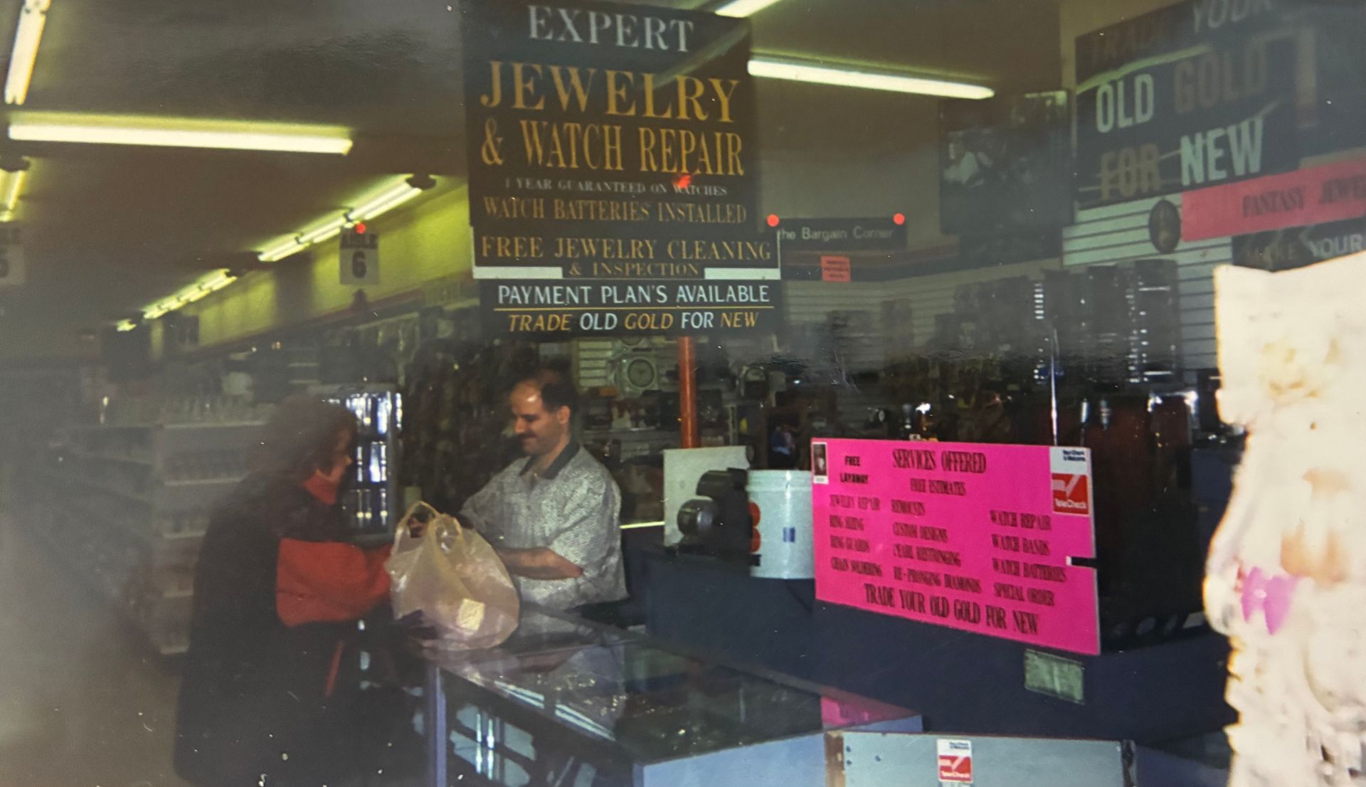 Two people inside a jewelry and watch repair shop, with gold and pink signage in the window.