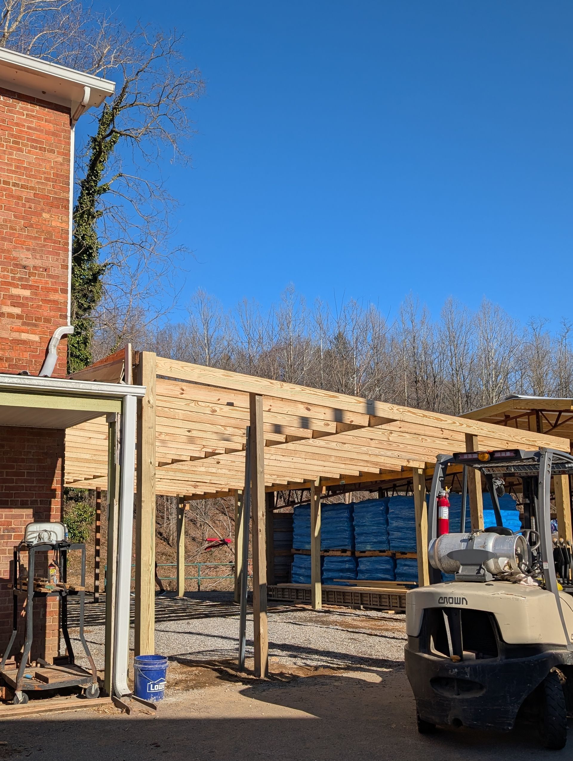 A wooden pergola under construction next to a brick building on a sunny day. A forklift is parked nearby.