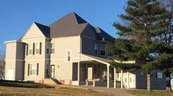 Large beige two-story house with gray roof and porch, viewed from the lawn under a clear blue sky