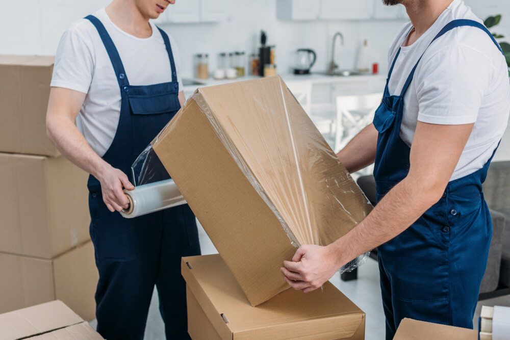 Two Men Are Wrapping a Cardboard Box With Plastic Wrap — A-Team Delivery Services Pty Ltd in Cessnock, NSW