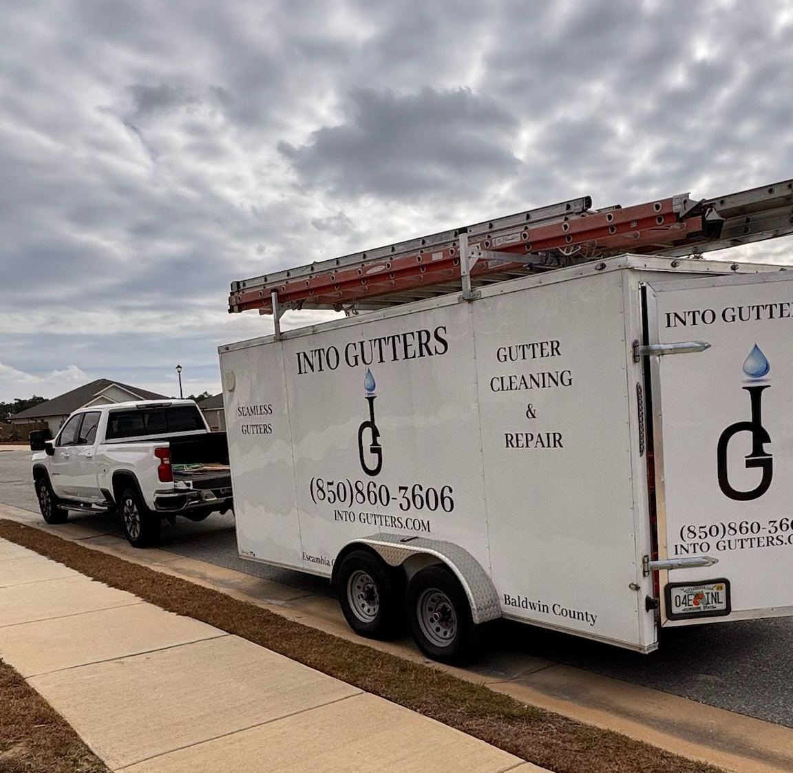 White pickup towing a branded moving trailer on a cloudy street beside a sidewalk