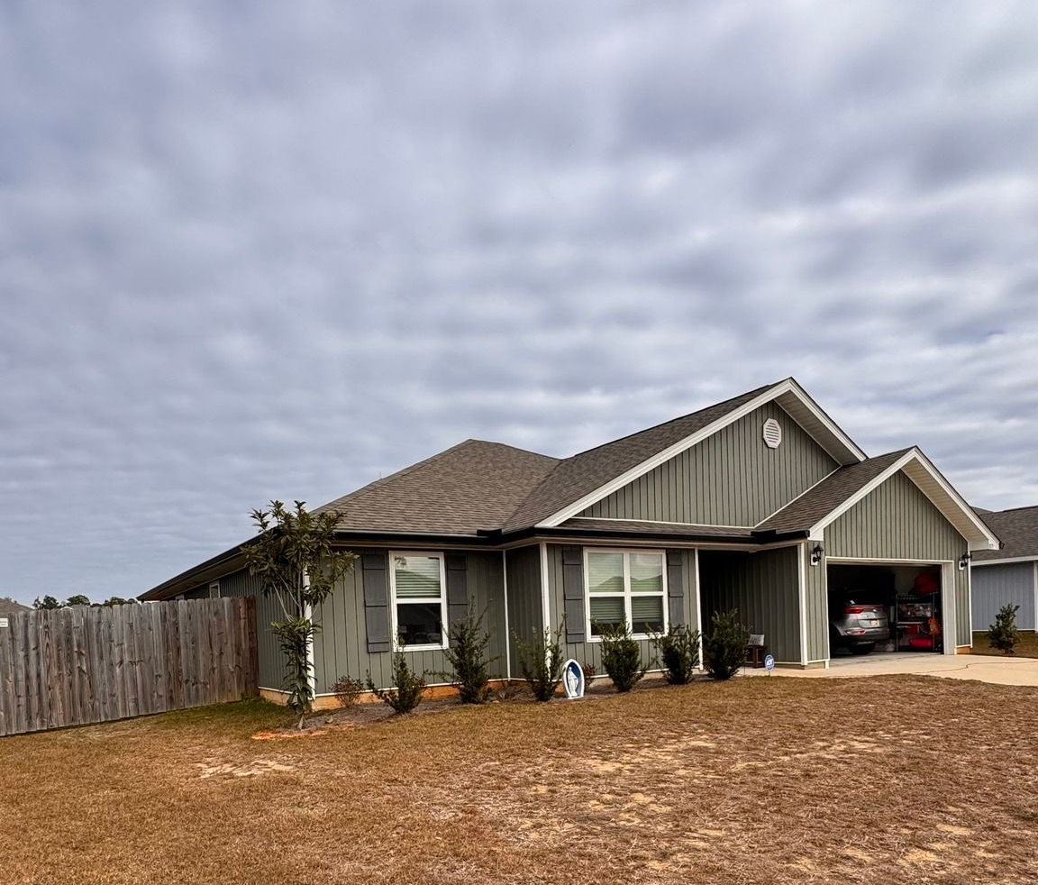 A single-story, grey-sided residential house with a shingled roof, front-facing garage, and a wooden fence on the left.