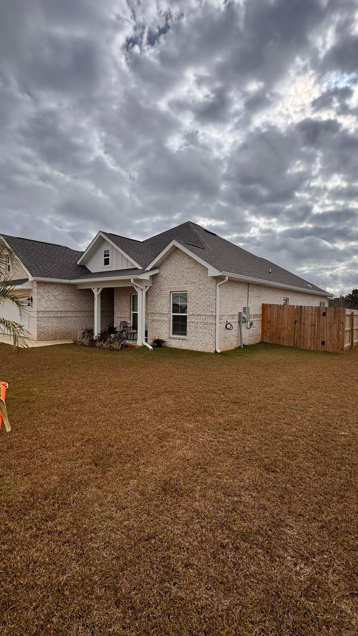 A single-story, light-colored brick house with a dark shingled roof under a cloudy, overcast sky.