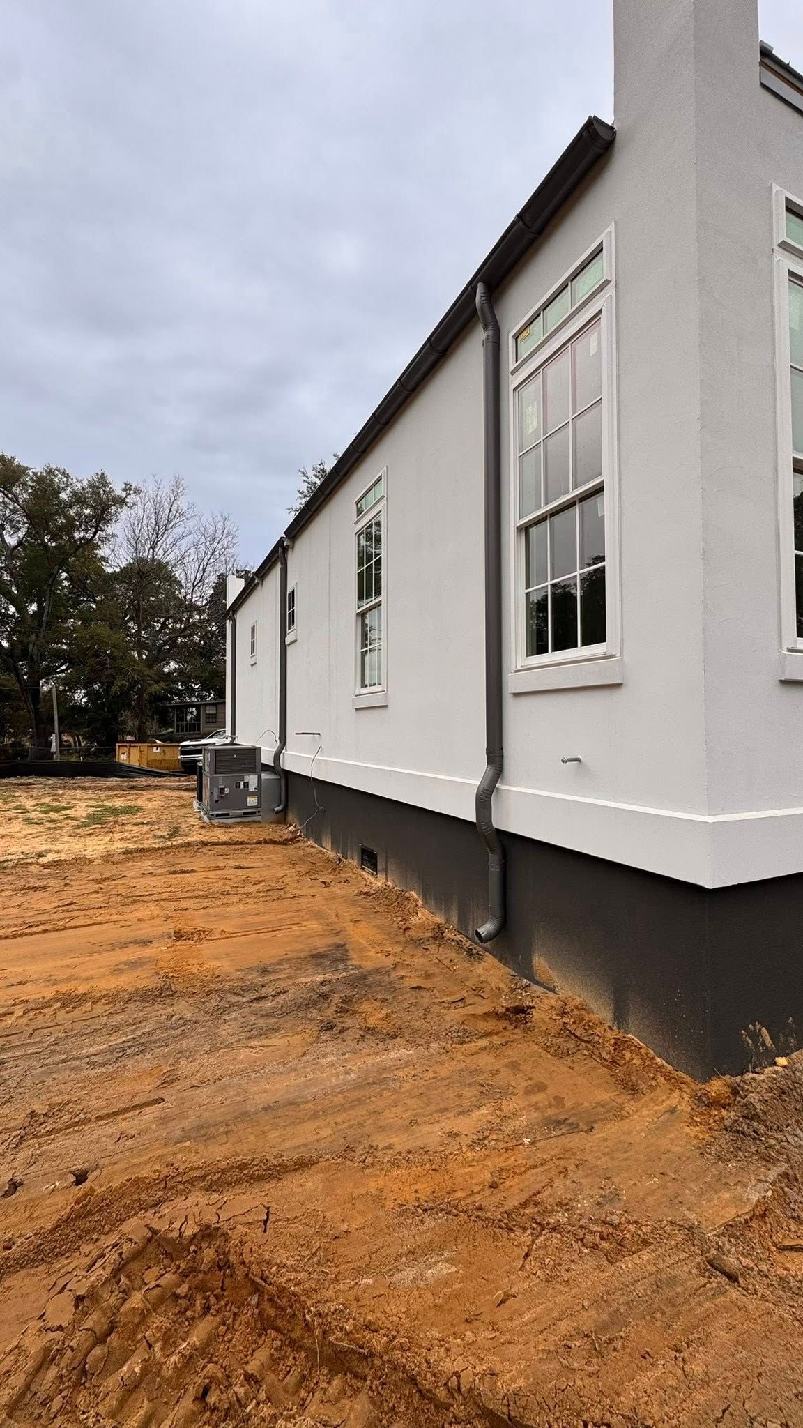 A light-colored house exterior with a dark foundation, tall windows, and downspouts over a dirt construction site.