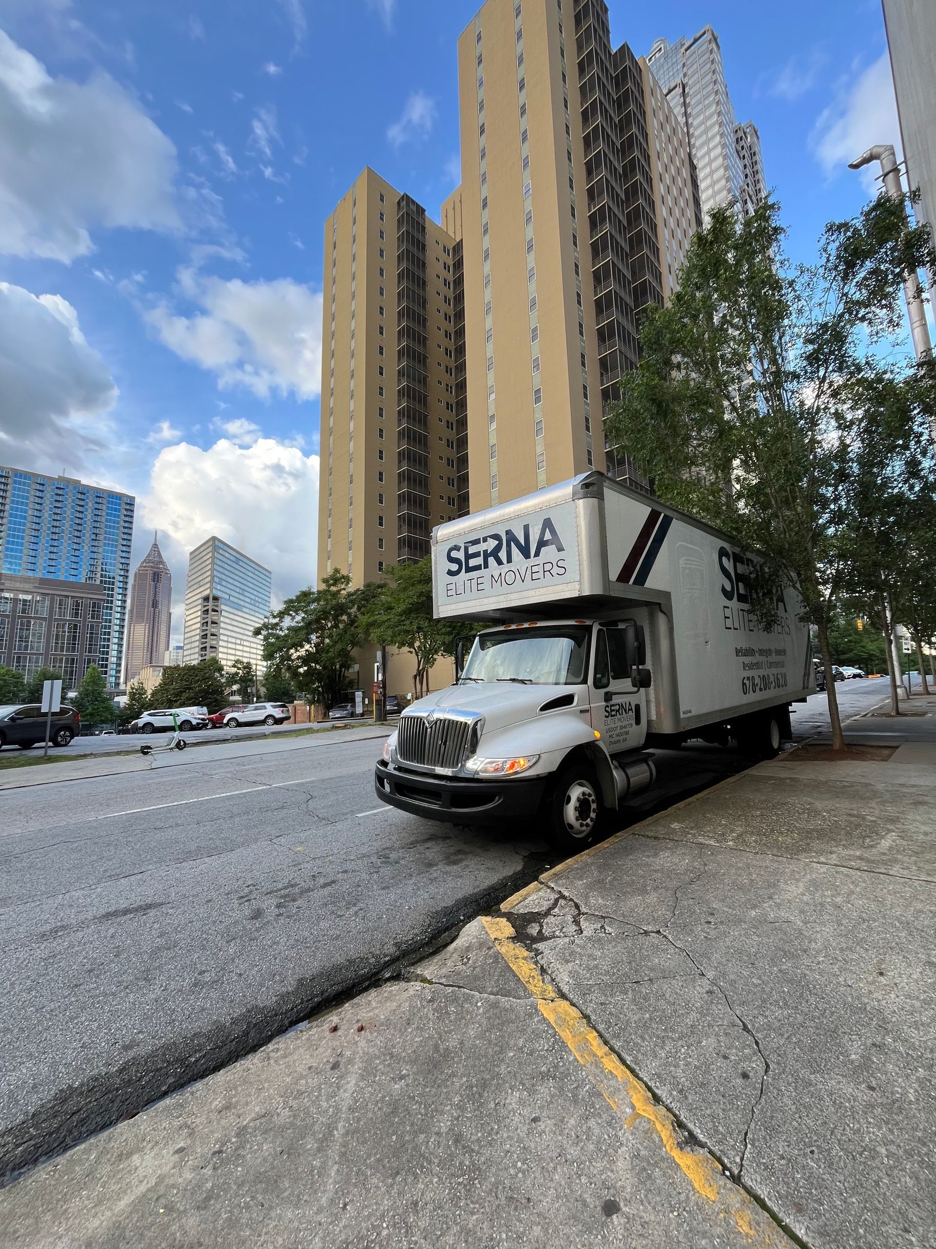 Moving truck parked on a city street, tall buildings in the background under a blue sky.