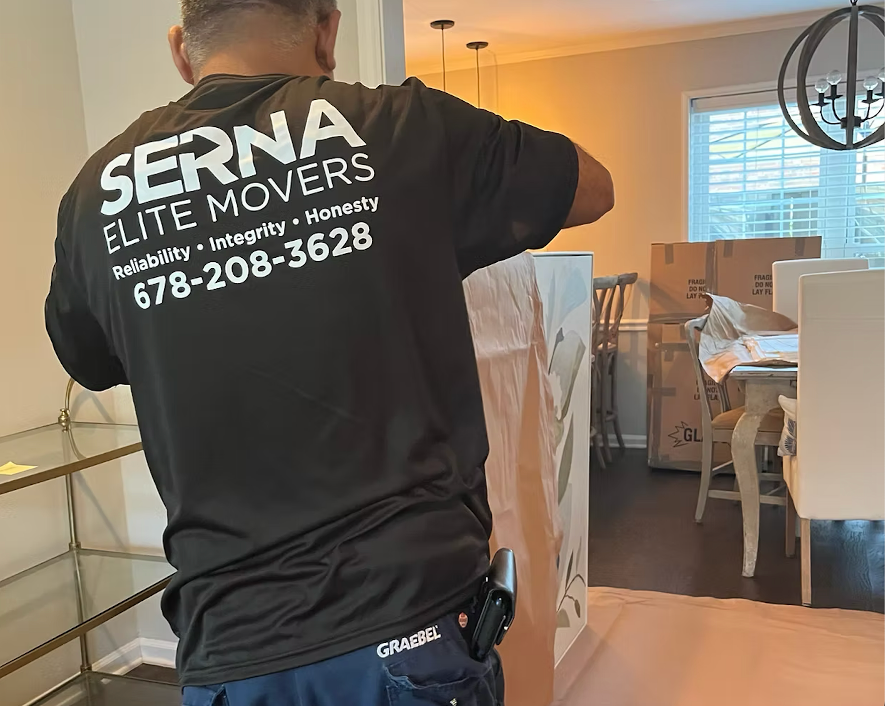 A man wearing a serna elite movers shirt is standing in a living room.