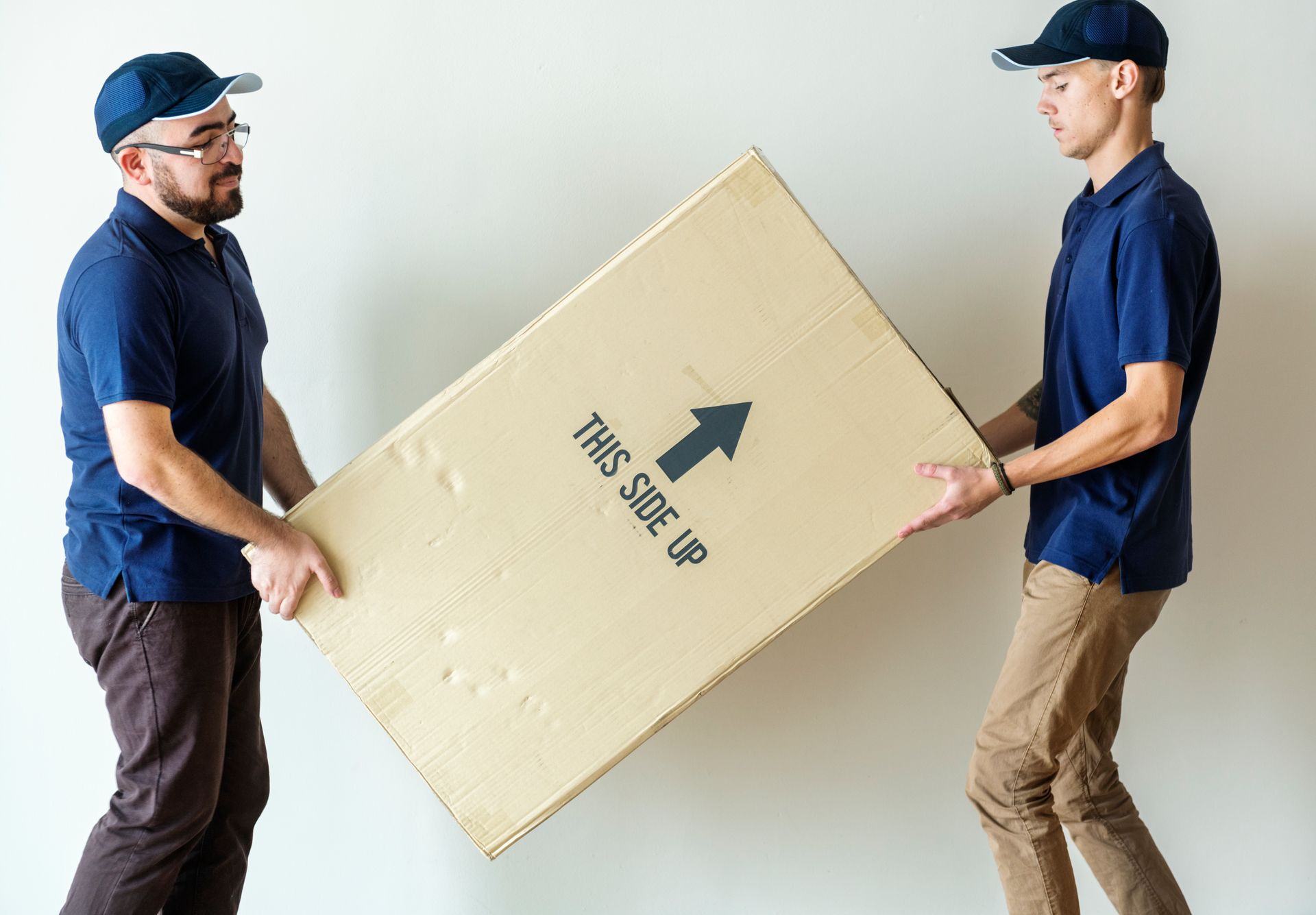 Two movers carrying a large cardboard box marked “This Side Up”