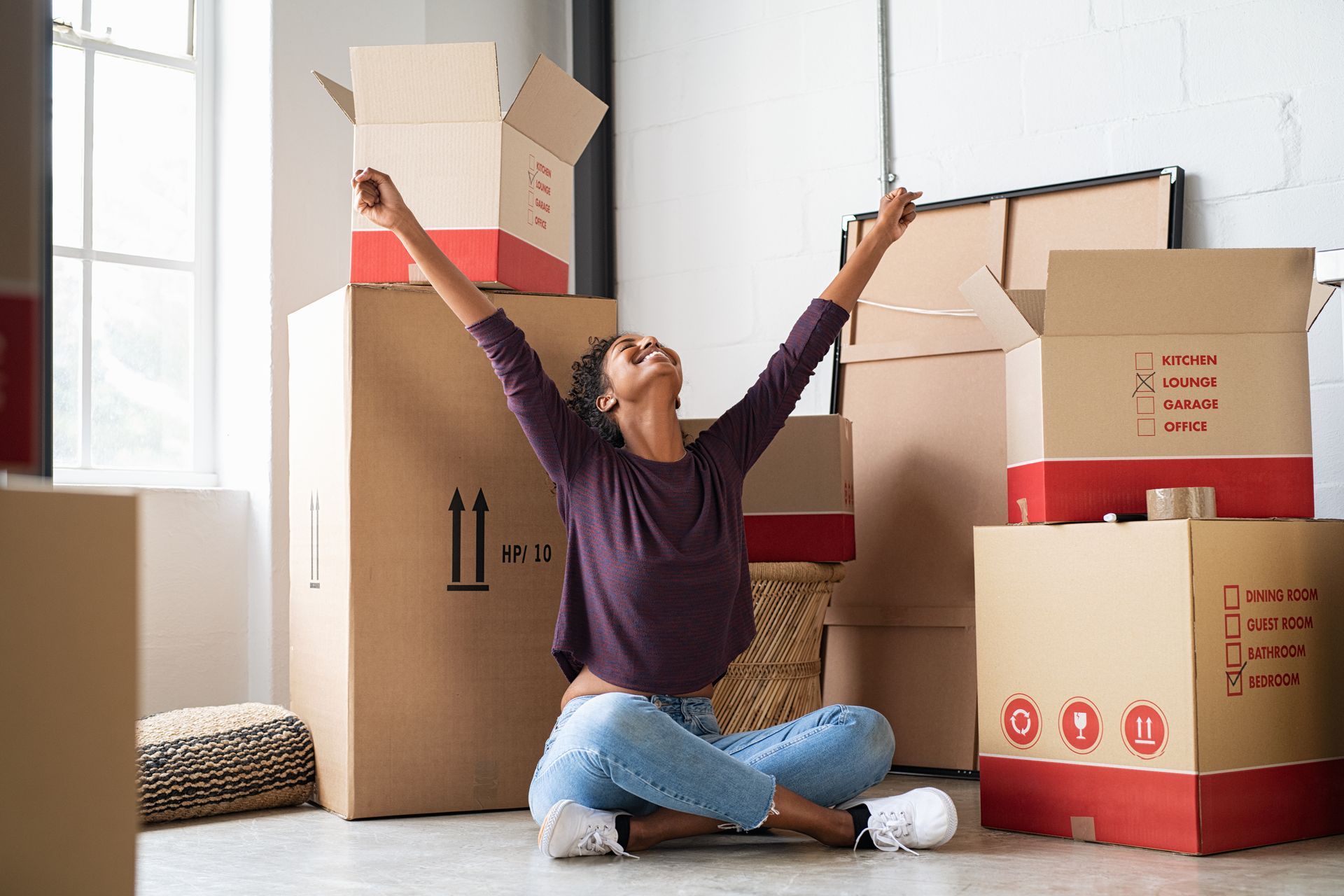 Woman in casual clothes celebrates in new home, surrounded by moving boxes.