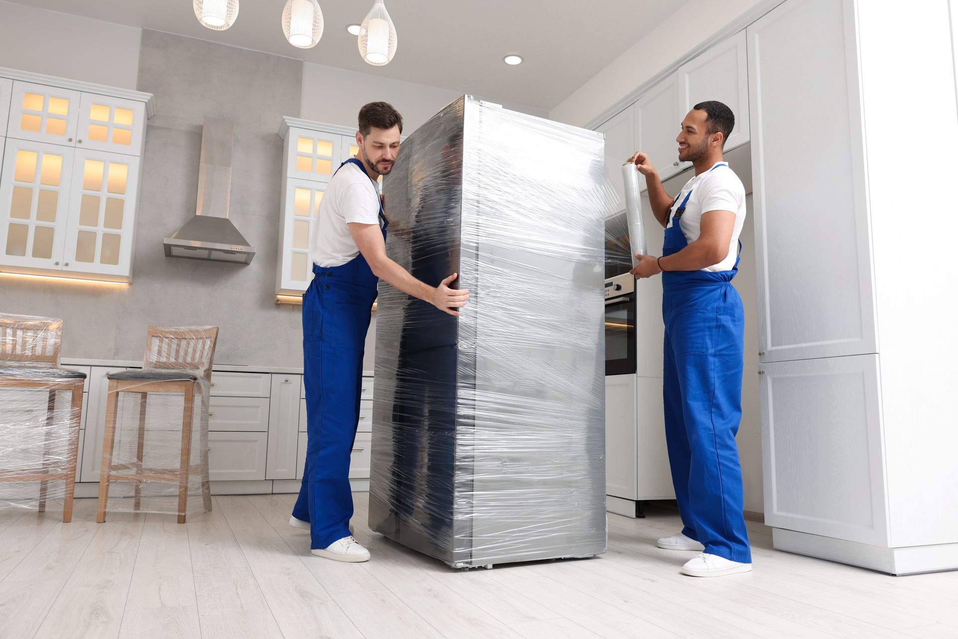 Two movers in blue uniforms carrying a wrapped refrigerator in a bright kitchen
