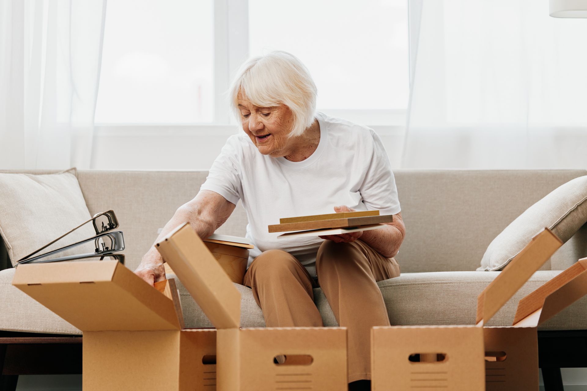 Elderly woman sorting items from boxes while sitting on a sofa. Indoors, natural light.