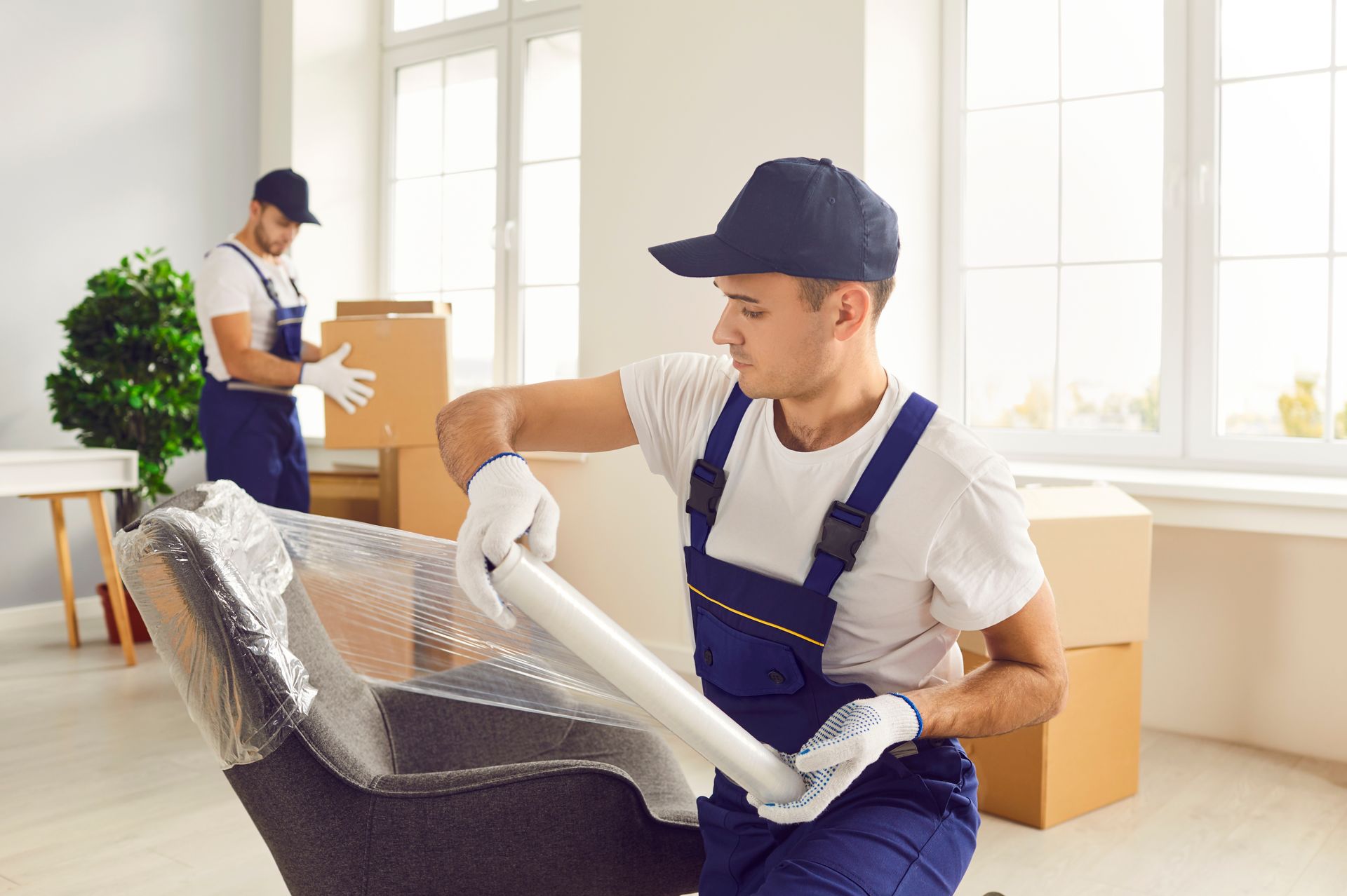 Two movers packing furniture with bubble wrap in a bright room with boxes and a window.