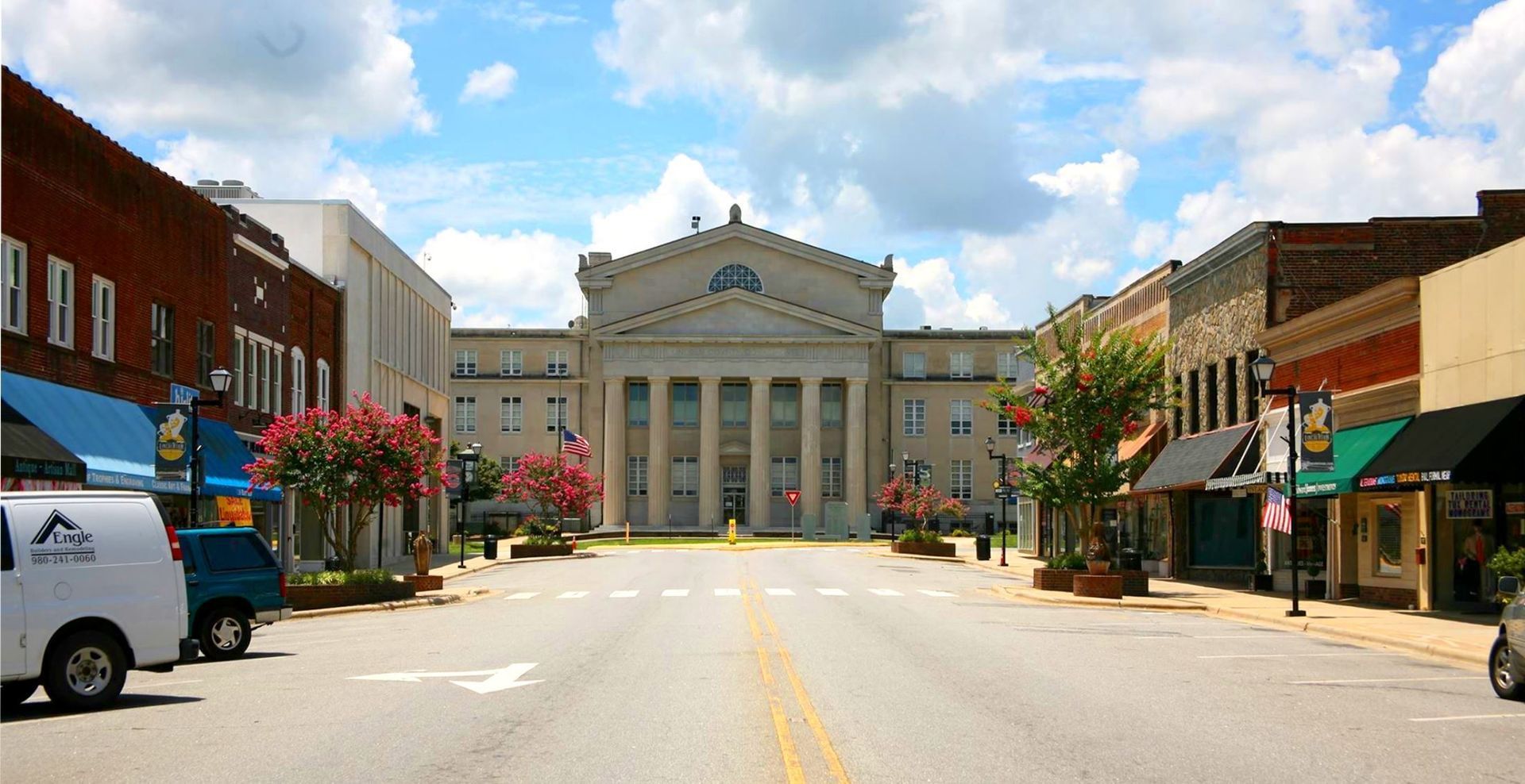 Small-town main street with brick storefronts leading to a white courthouse under a blue sky