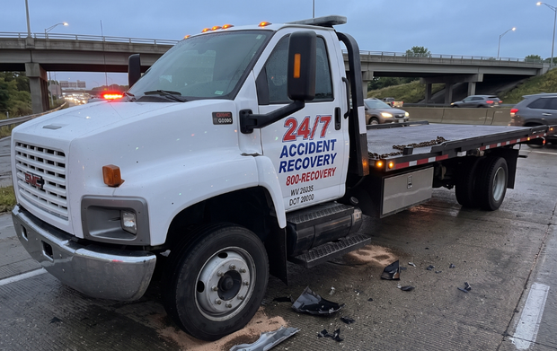 White tow truck on wet highway shoulder after crash, with debris scattered beside an overpass.