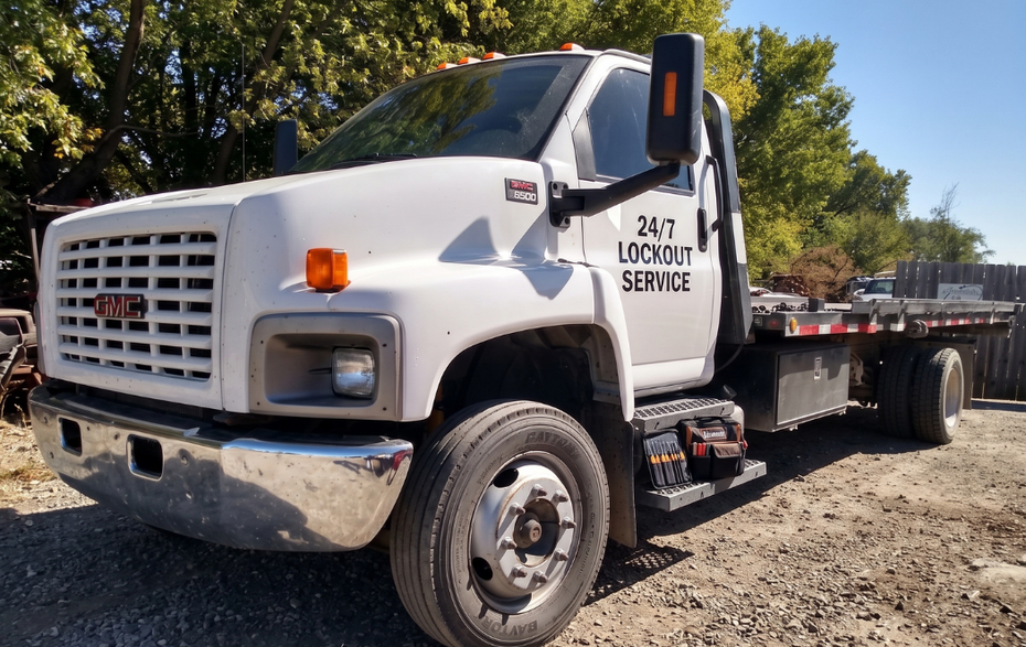 White flatbed tow truck parked on gravel under trees, side view with company logo on the door