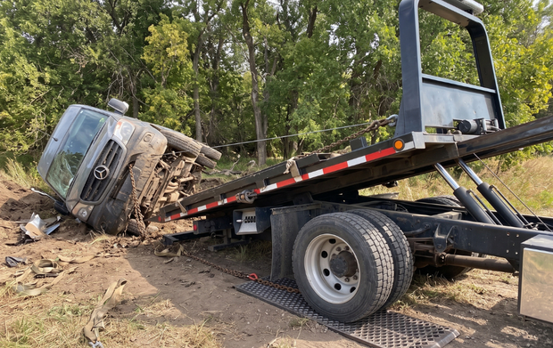 Tow truck pulling a muddy overturned truck off a dirt roadside in a wooded area