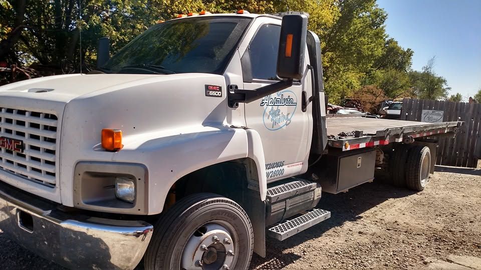 White flatbed tow truck parked on a gravel lot beside trees in bright sunlight