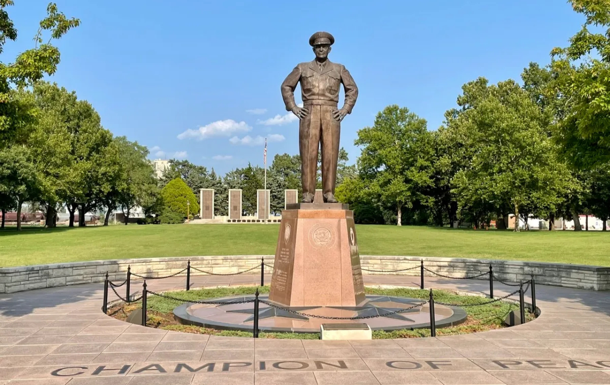 Bronze statue on a stone pedestal in a sunny park, surrounded by trees and a circular walkway