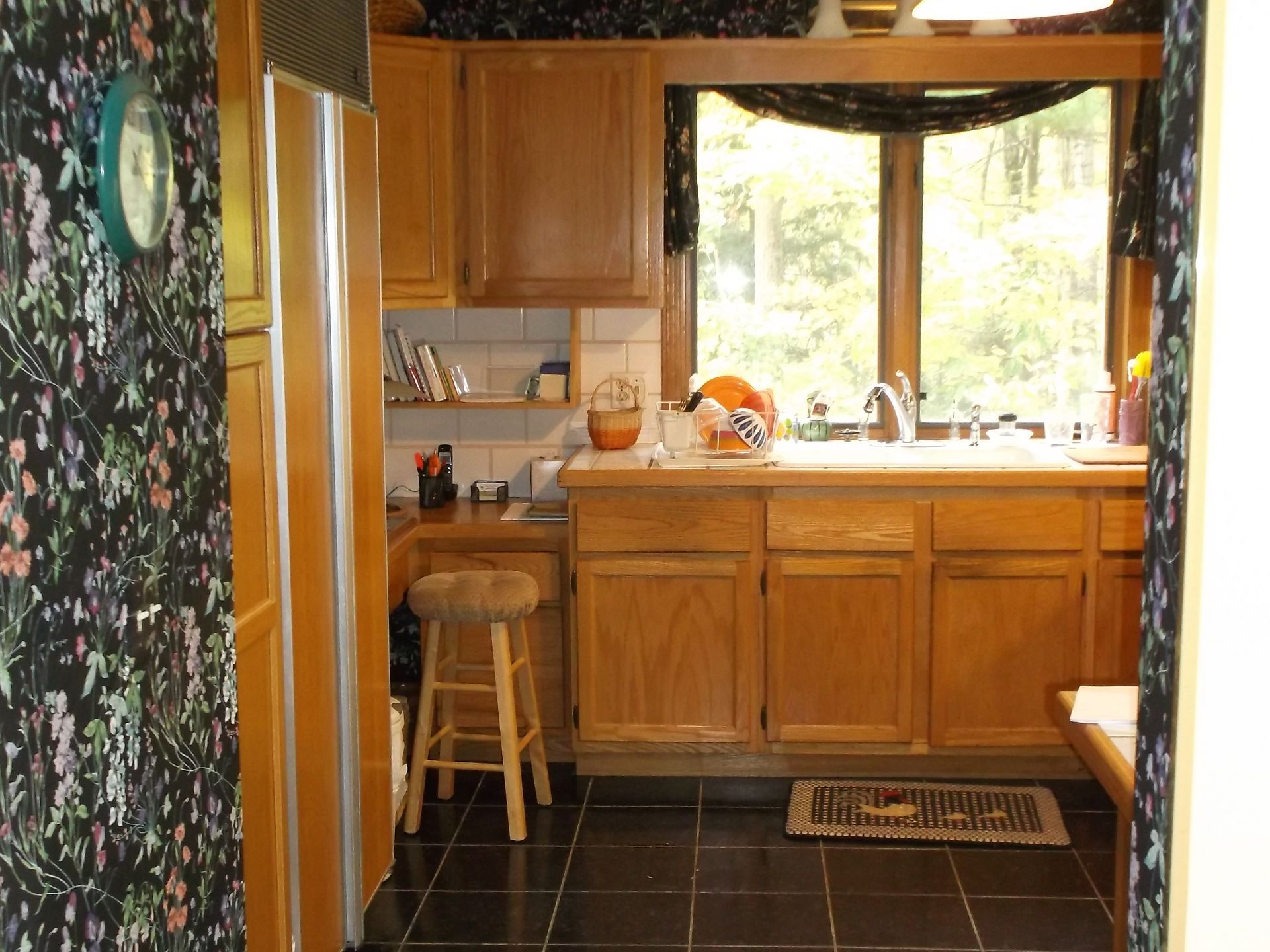 Warm kitchen with wooden cabinets, floral curtains, a stool by the counter, and sunlight through a window.