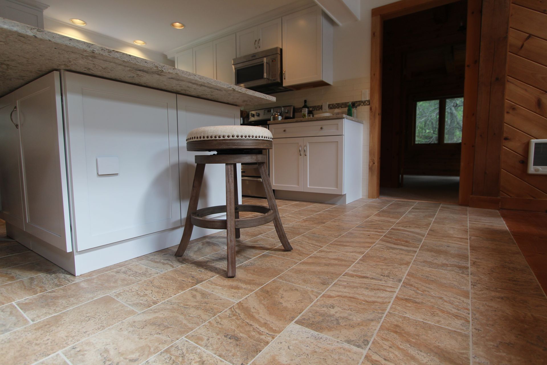 Modern kitchen with white cabinets, stone tile floor, central stool, and dark wood doorway