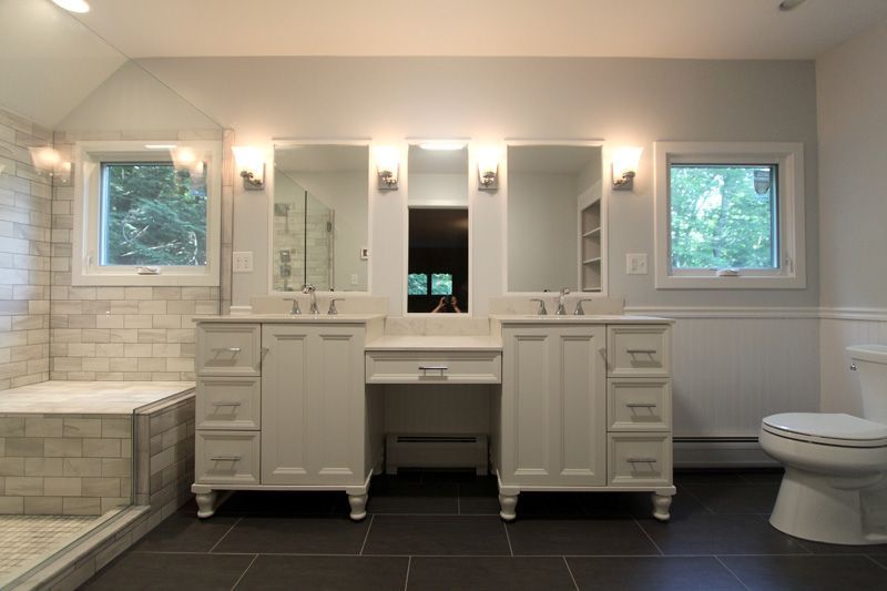 Bright bathroom with double vanity, mirrors, windows, bathtub, and toilet on dark tile floor