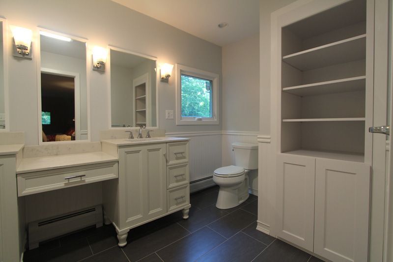 Bright bathroom with double vanity, mirrors, toilet, and built-in shelves, lit by wall sconces.