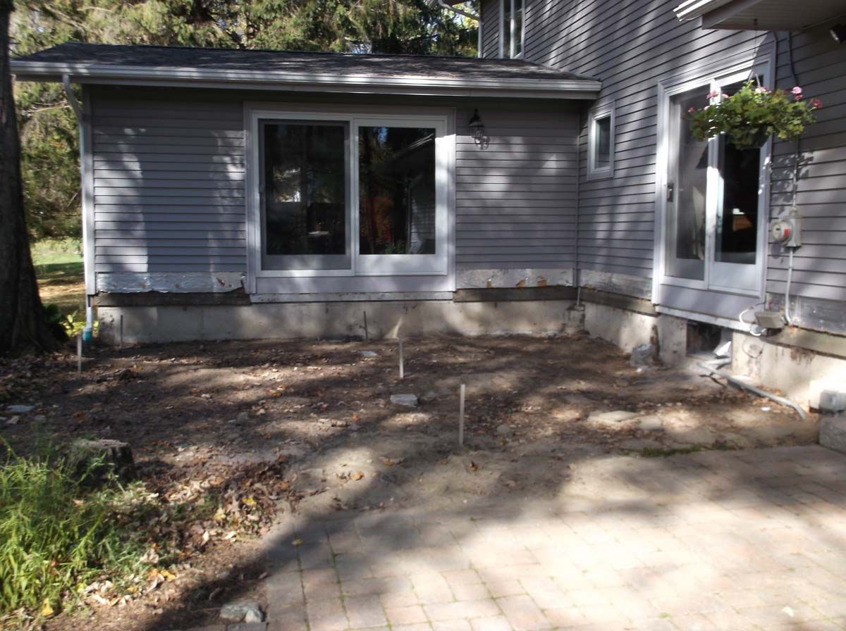 Backyard patio beside a gray house with windows and bare ground under trees