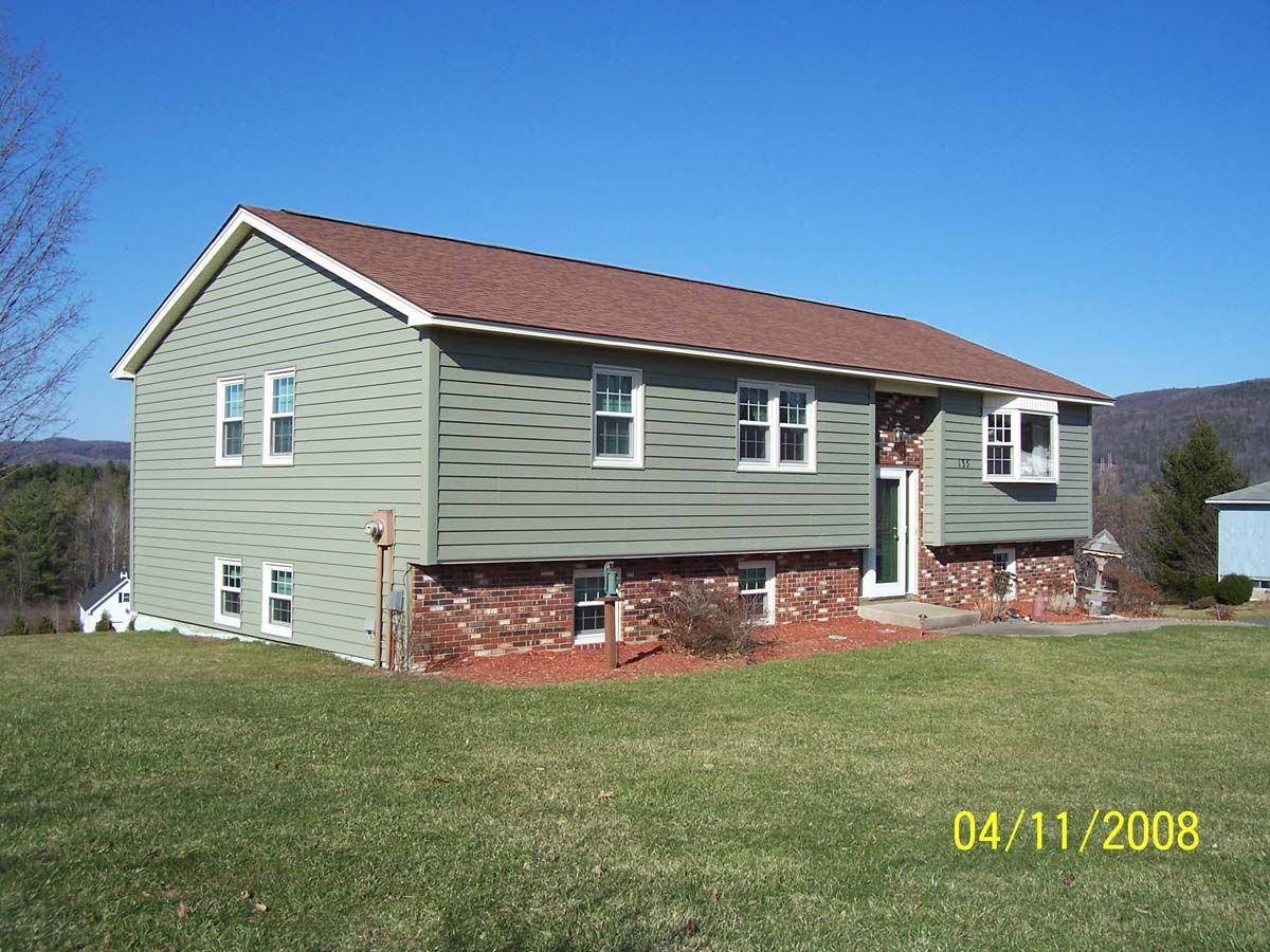 Side view of a light green two-story house with a red roof and brick foundation on a grassy lawn.
