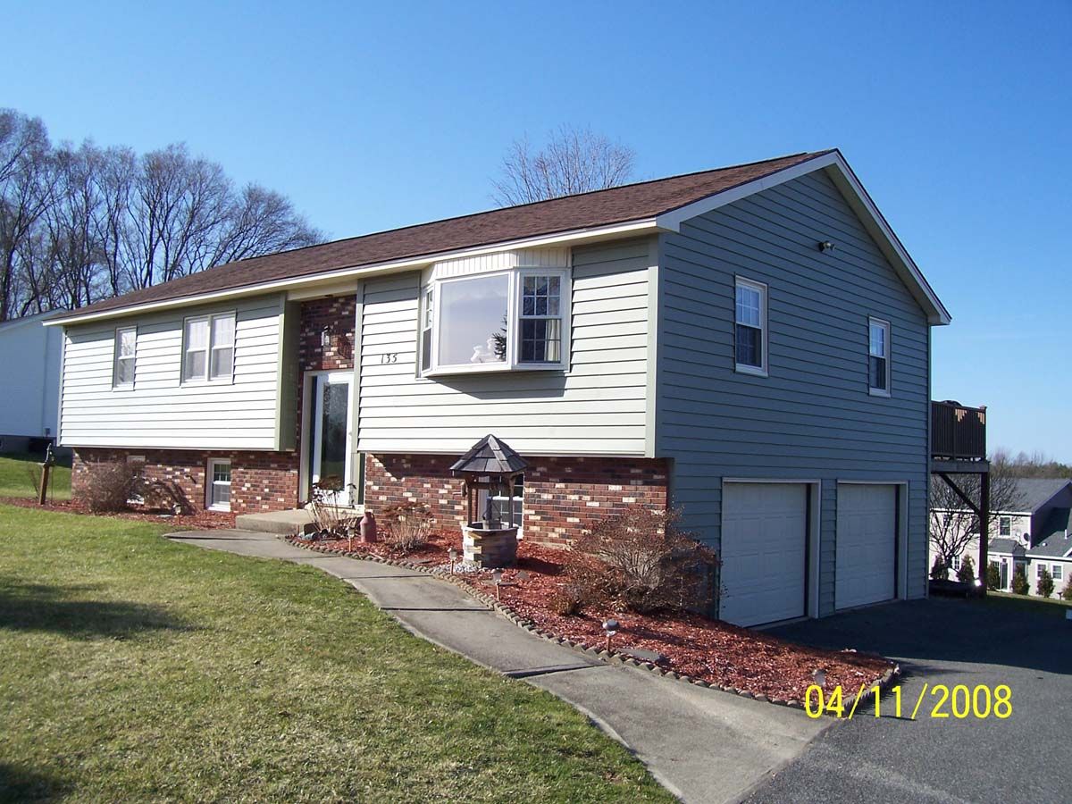 Suburban split-level house with beige siding, gray garage, red roof, and a front walkway on a sunny day