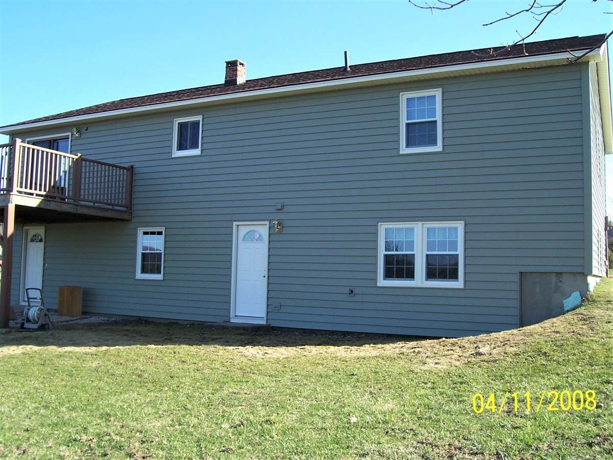 Two-story gray house with white trim, side deck, and grassy yard under a clear sky