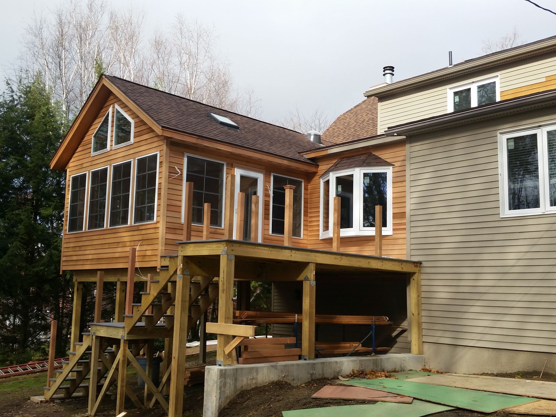 Wooden house extension on stilts beside a brick home, with a deck and unfinished yard below.